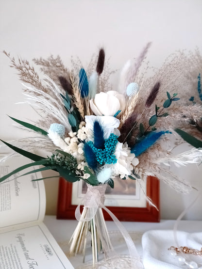 This handcrafted wedding bouquet features an exquisite blend of dried flowers, combining soft natural tones with striking blue accents. The arrangement includes pampas grass, preserved foliage, and delicate bunny tails, creating a perfect balance of rustic charm and modern elegance. Ideal for a boho-themed wedding, this long-lasting bouquet adds a touch of timeless beauty to your special day. Explore our collection for more unique bridal bouquets and wedding decor ideas.