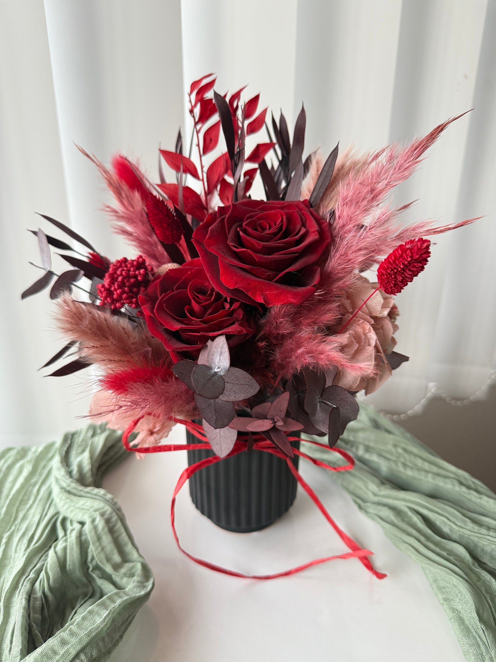 Bouquet of red roses and pink flowers in a black vase on a white surface with a green cloth.