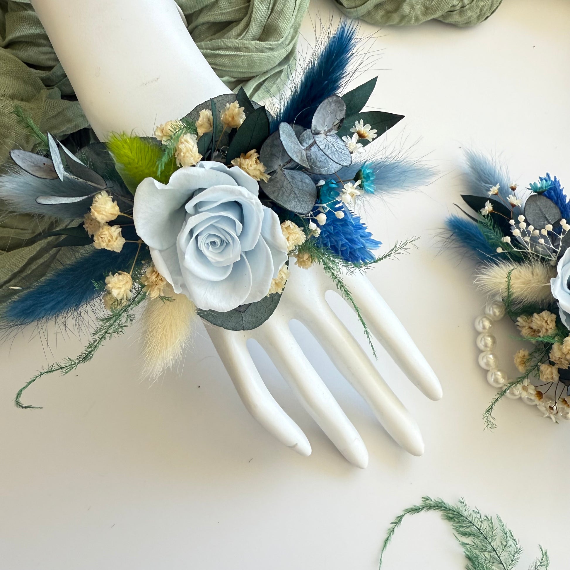 Handmade wrist corsage with dusty blue rose, ivory flowers, and greenery on mannequin hand.
