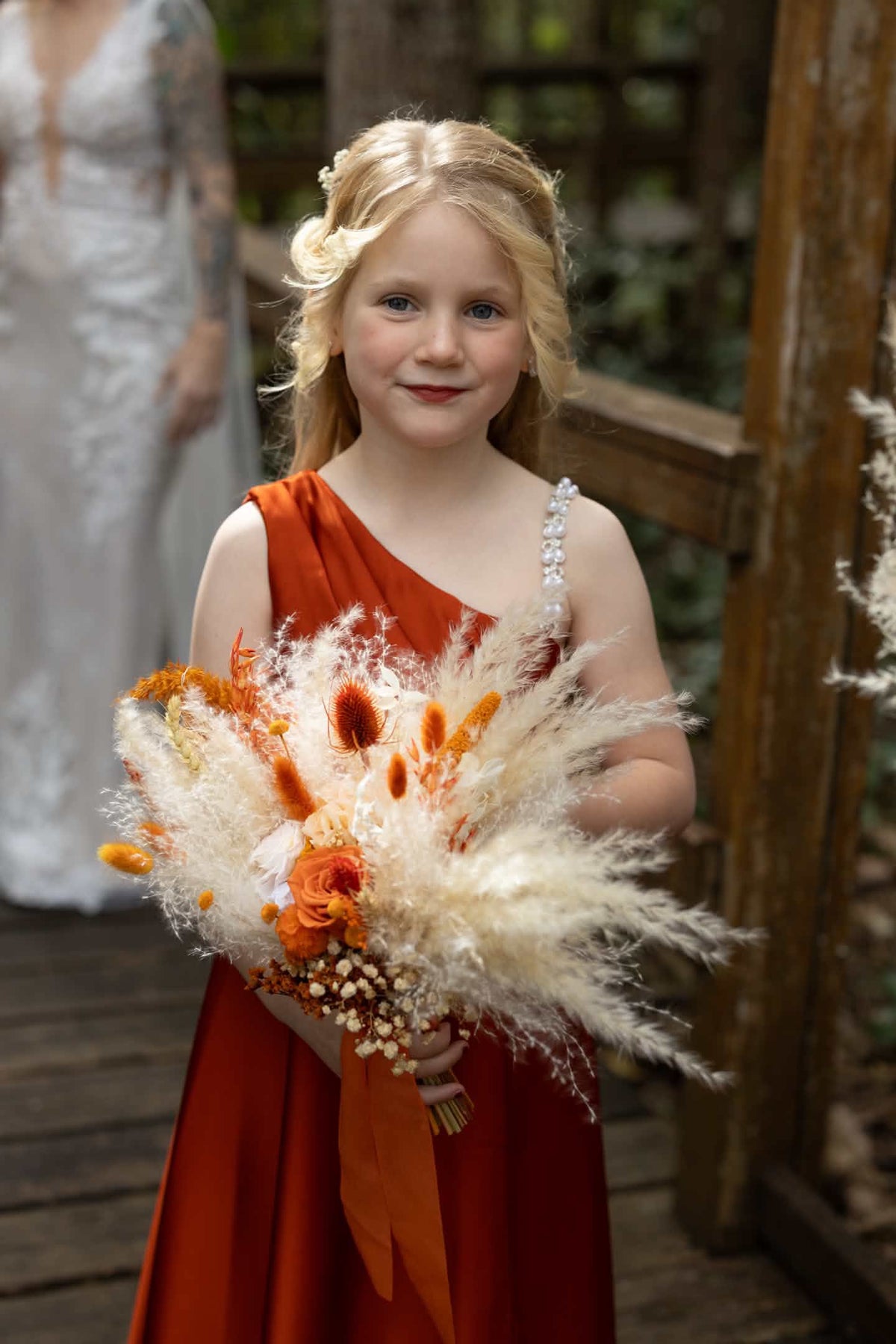 A young girl with blonde hair stands outdoors holding a large autumn bouquet with orange flowers and beige pampas grass. She is wearing a rust-orange one-shoulder dress, with a blurred person in a white dress in the background.