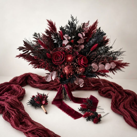 A dark red and black floral bouquet arrangement with burgundy ribbon, displayed on a light neutral background with matching accessory pieces and a draped burgundy fabric.