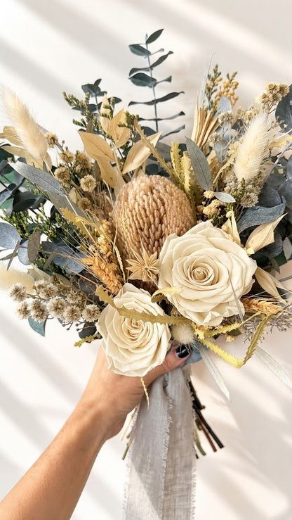 A dried floral bouquet held by a hand, featuring cream roses, a large protea, pampas grass, and green foliage against a white background with soft window shadows.