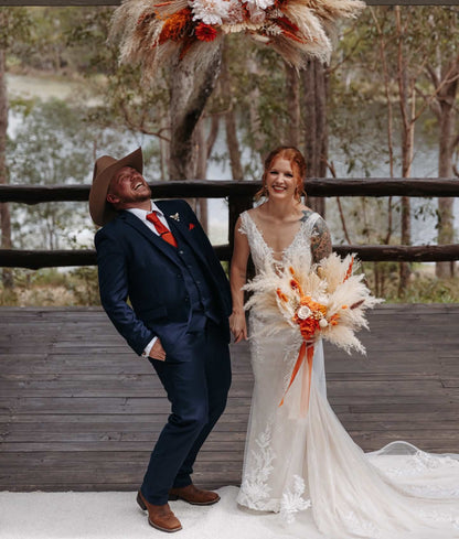 A bride in a white lace wedding gown holding an orange and beige pampas flower bouquet stands beside a groom in a navy blue suit and brown cowboy hat, posing on a wooden deck with a large pampas flower arch overhead.