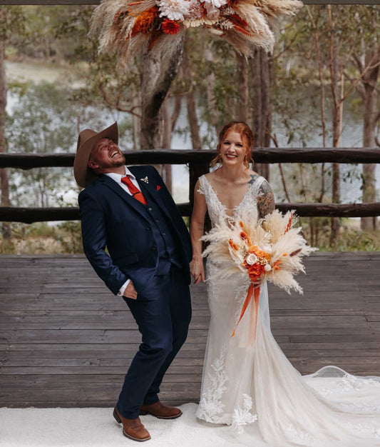 A bride in a white lace wedding gown holding an orange and beige pampas flower bouquet stands beside a groom in a navy blue suit and brown cowboy hat, posing on a wooden deck with a large pampas flower arch overhead.