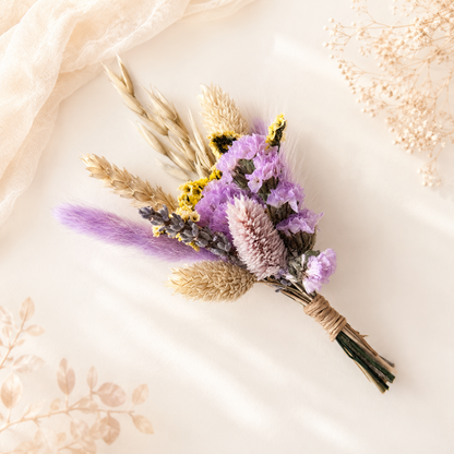A dried floral bouquet with purple flowers, beige wheat stalks, and a fluffy lavender tail, wrapped in twine and photographed on a soft cream fabric background with decorative dried botanicals.