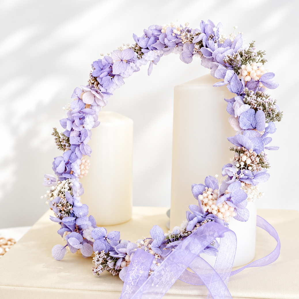 A lavender floral wreath with purple hydrangeas, white baby's breath, and a matching purple ribbon bow, displayed on cream books and candles.