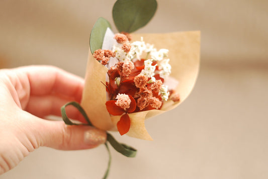 Mini dried flower bouquet in warm terracotta and white tones, held in hand. Wrapped in kraft paper with a green ribbon and a touch of eucalyptus leaf. Includes dried baby’s breath and strawflowers. Perfect for rustic weddings, small thank-you gifts, or eco-friendly event favours. Soft natural lighting creates a cozy and handmade aesthetic.