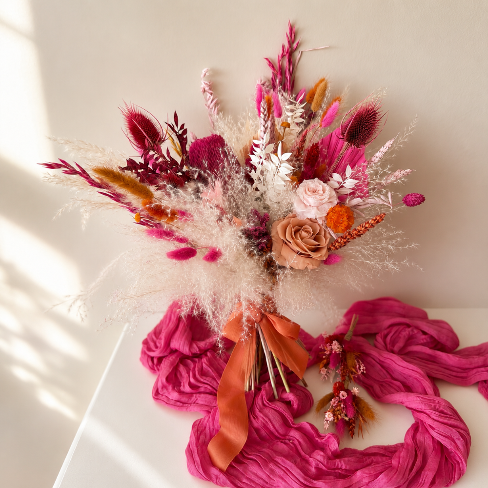 Large dried flower bridal bouquet with preserved blush roses, pink pampas grass, banksia, bunny tails, and orange accents styled with flowing pink silk ribbon on a light background.