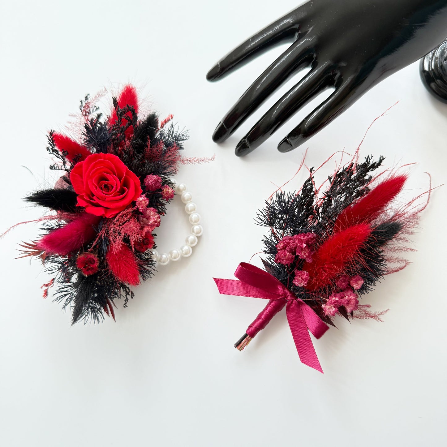 Red and black dried boutonniere with preserved rose and matching corsage displayed together on white background.
