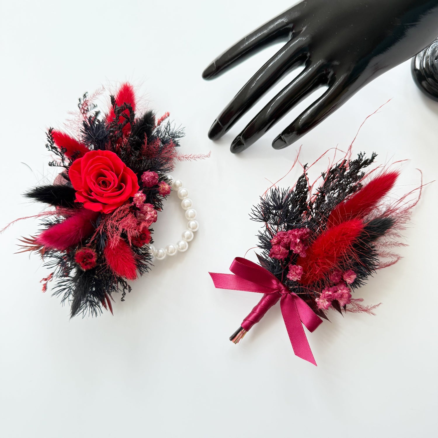 Red and black dried boutonniere with preserved rose and matching corsage displayed together on white background.