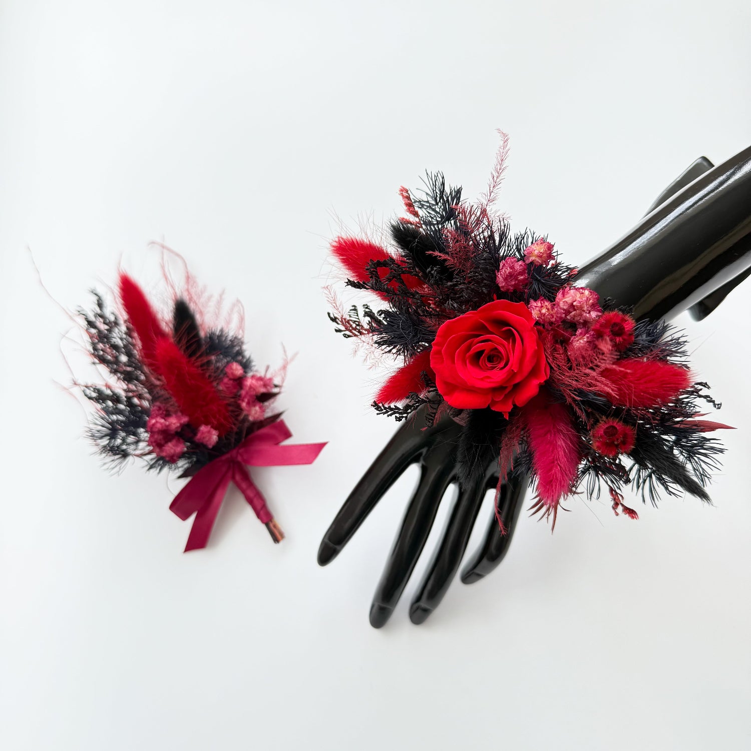 Red and black dried corsage and boutonniere set with preserved roses on black display hand.