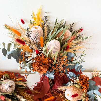 Large rustic bridal bouquet with banksia, eucalyptus, hydrangea, bunny tails, and burnt orange dried flowers, styled with a deep red ribbon.