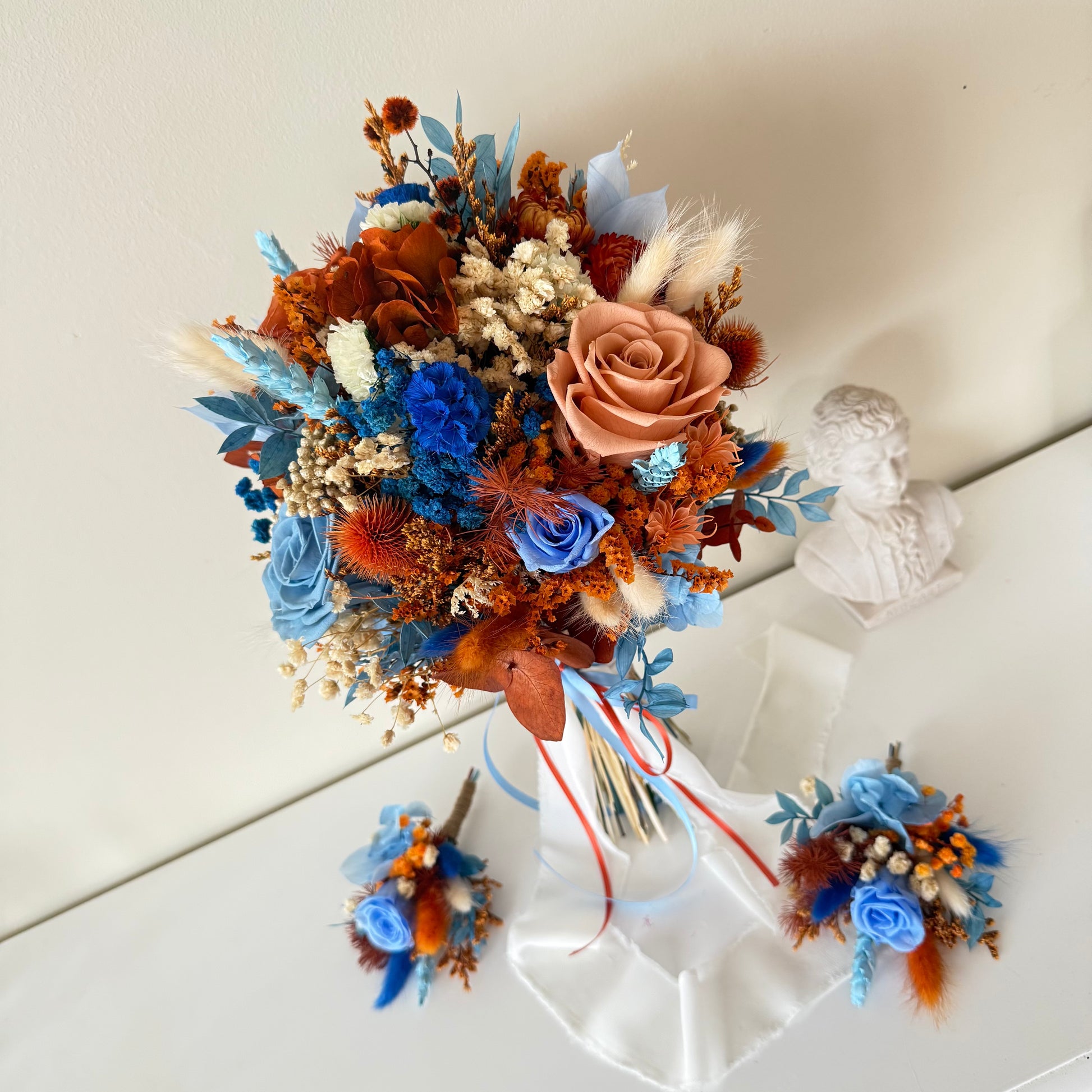 This beautifully crafted wedding bouquet features an elegant mix of orange, white, and natural tones, made from dried flowers and pampas grass. The arrangement exudes a modern rustic charm, with preserved foliage and delicate accents adding a sophisticated touch. Perfect for a contemporary bride, this long-lasting bouquet is ideal for weddings with a natural or bohemian theme. Explore our collection for more unique bridal bouquets and wedding decor inspirations