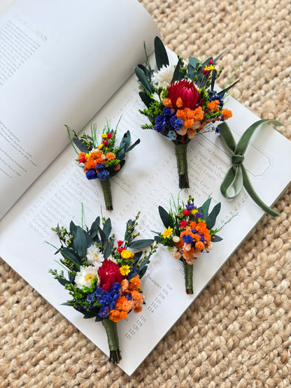 Rustic dried flower boutonniÃ¨re set with burnt orange, royal blue and emerald green tones on neutral background.