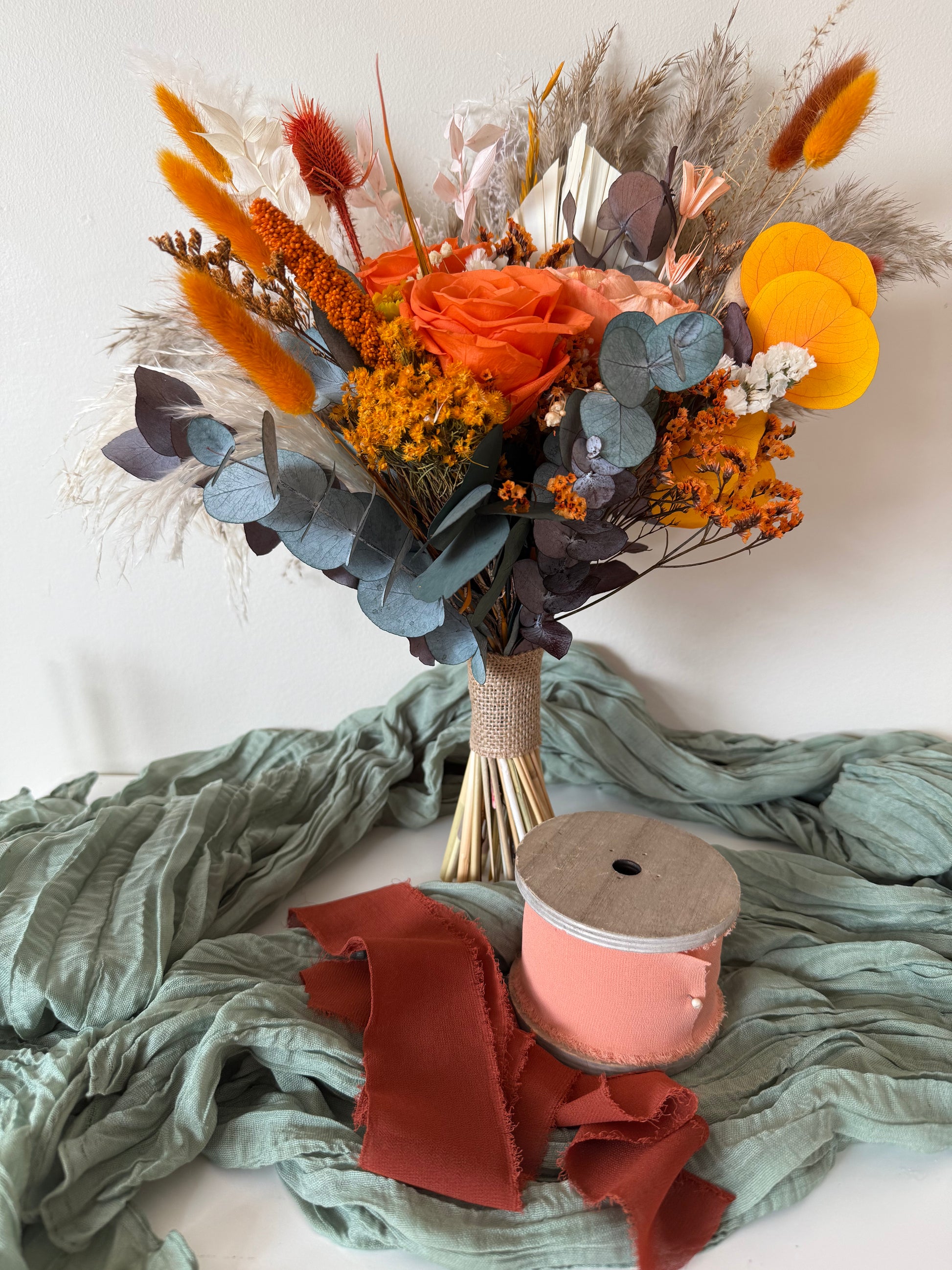 Boho bridal bouquet with terracotta, orange, and yellow dried flowers, sola wood roses, pampas grass, and eucalyptus, tied with a dusty pink ribbon, styled with green fabric.