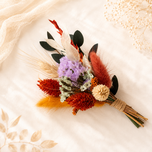 A bohemian-style dried flower bouquet with purple flowers, red-orange grasses, beige feathers, and green leaves, wrapped in twine and laid on a soft cream fabric background.
