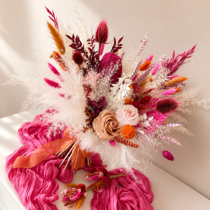 Boho wedding bouquet featuring preserved roses, pink banksia, fluffy white pampas grass, orange dried flowers, and bright pink silk styling fabric arranged for bridal inspiration