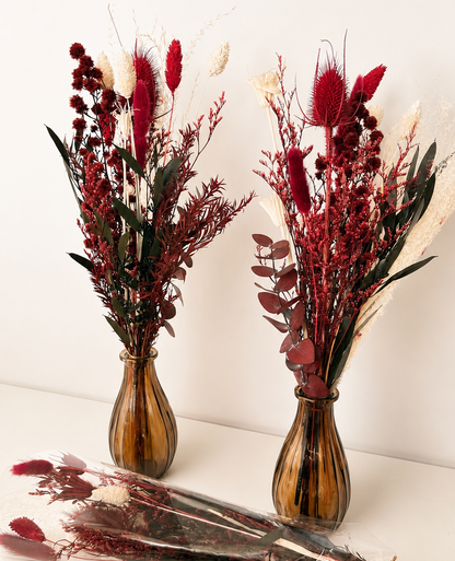 Two amber glass vases filled with dried decorative flowers in deep red, burgundy, and cream tones, arranged on a white surface against a plain light background. Several wrapped bundles of similar dried stems lie in front.