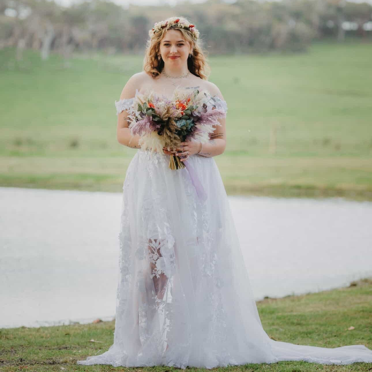 Bride standing outdoors near a lakeside during an Australian wedding, holding a large boho dried flower bouquet in pastel colours
