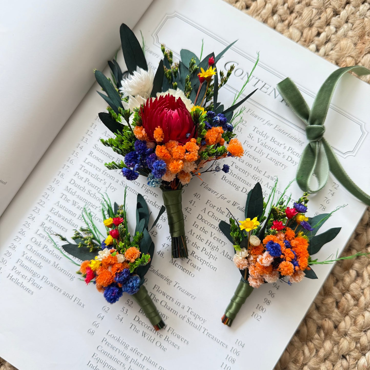 Australian dried flower boutonniÃ¨res featuring red protea, orange broom bloom and blue statice on book background.