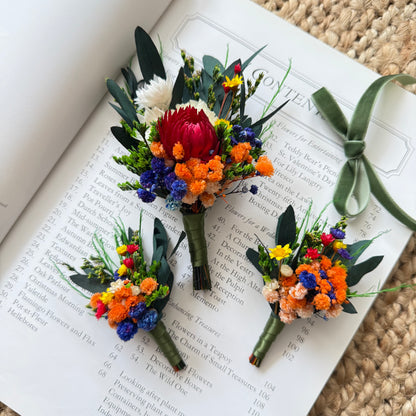 Australian dried flower boutonniÃ¨res featuring red protea, orange broom bloom and blue statice on book background.