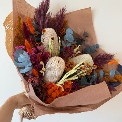 Colourful Australian native dried flower bouquet wrapped in brown kraft paper with banksia, eucalyptus and orange and burgundy blooms.