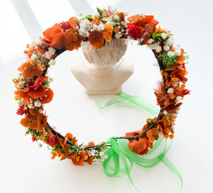 An autumn floral wreath with orange and red dried flowers, white filler blossoms, green stems, and a sheer green ribbon bow, displayed on a white bust stand against a bright white background.