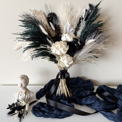 A dried floral arrangement featuring pampas grass in black, white, and dark green tones, with cream-colored flowers and a black ribbon, displayed on a white surface next to a small white bust and draped navy fabric.