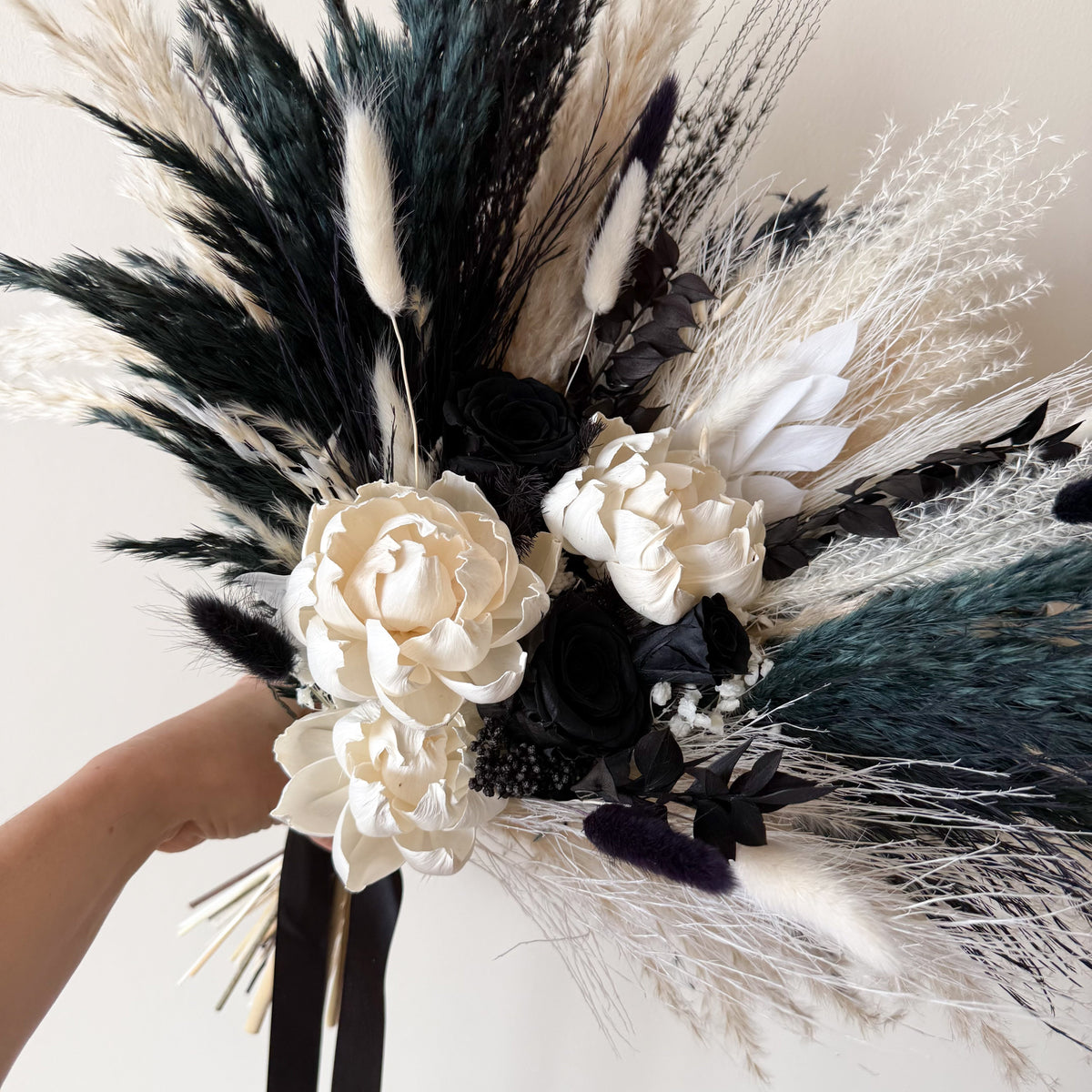 A dried floral bouquet held by a hand, featuring cream peony-like flowers, black roses, pampas grass in cream and dark teal, and delicate white filler stems against a plain light background.