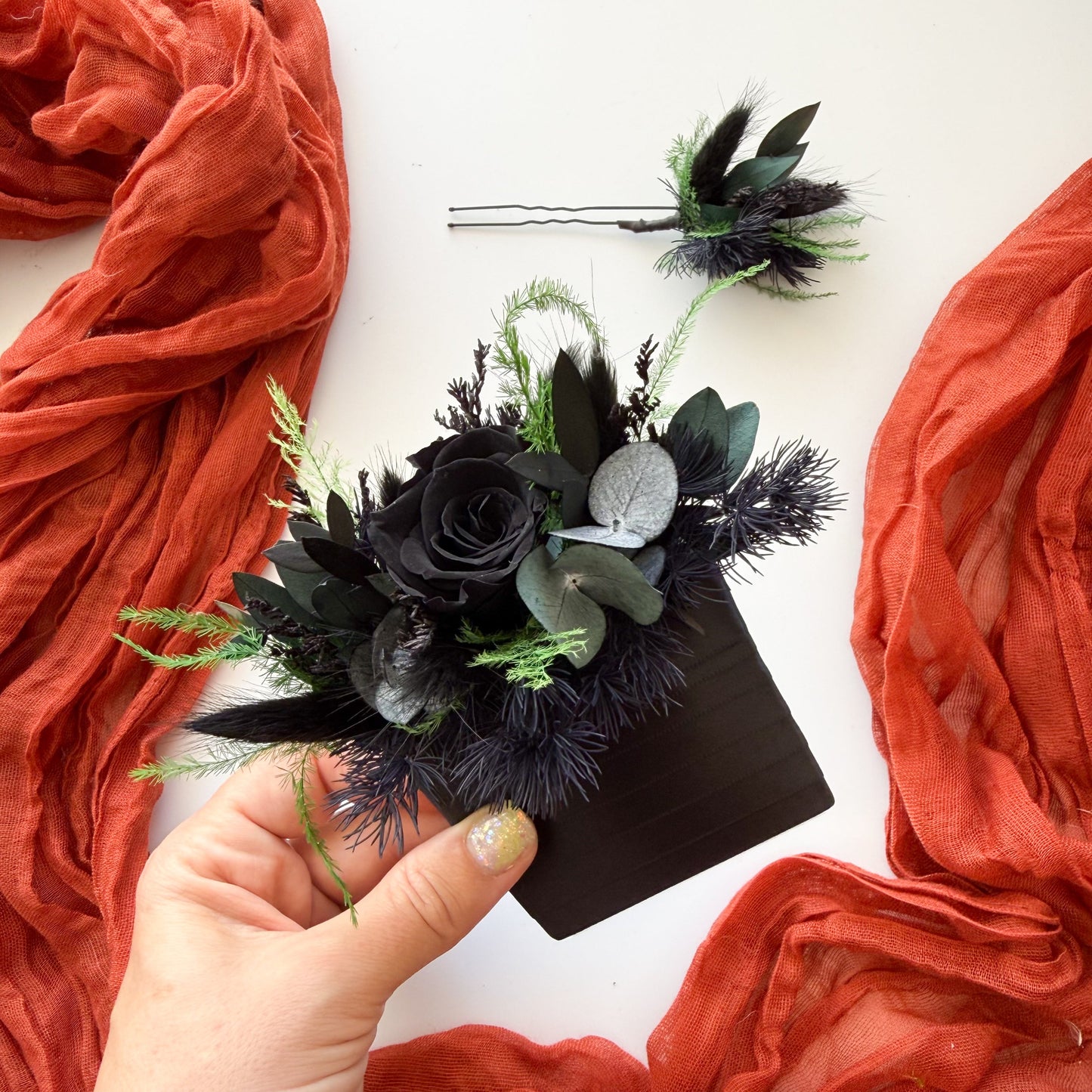 Hand holding a black boutonniere with greenery against an orange fabric background
