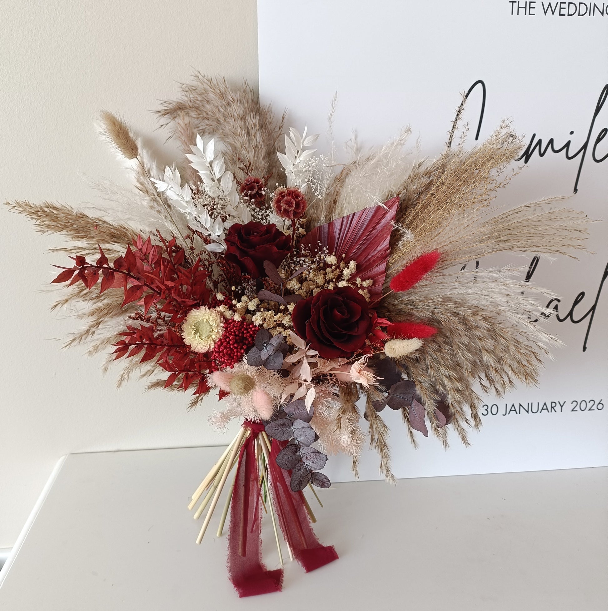 Boho bridal bouquet with dried flowers, pampas grass and dark red accents, styled next to a welcome sign.