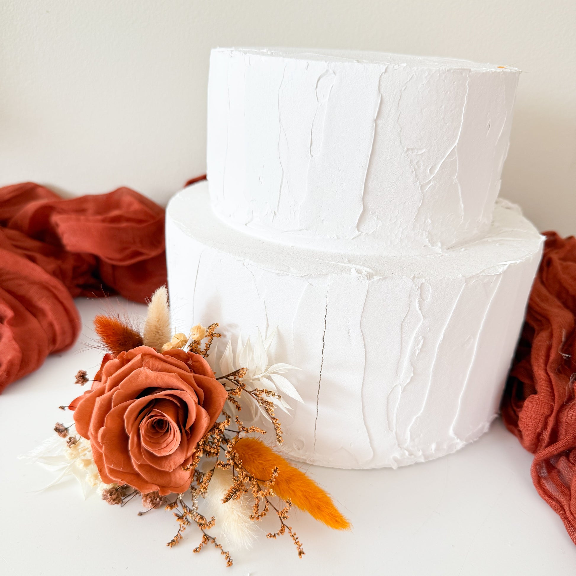 Terracotta dried floral cake decoration on two-tier white cake with rust fabric background