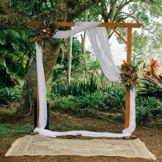 Boho outdoor wedding arch decorated with dried flower arrangements in warm tones and flowing white fabric
