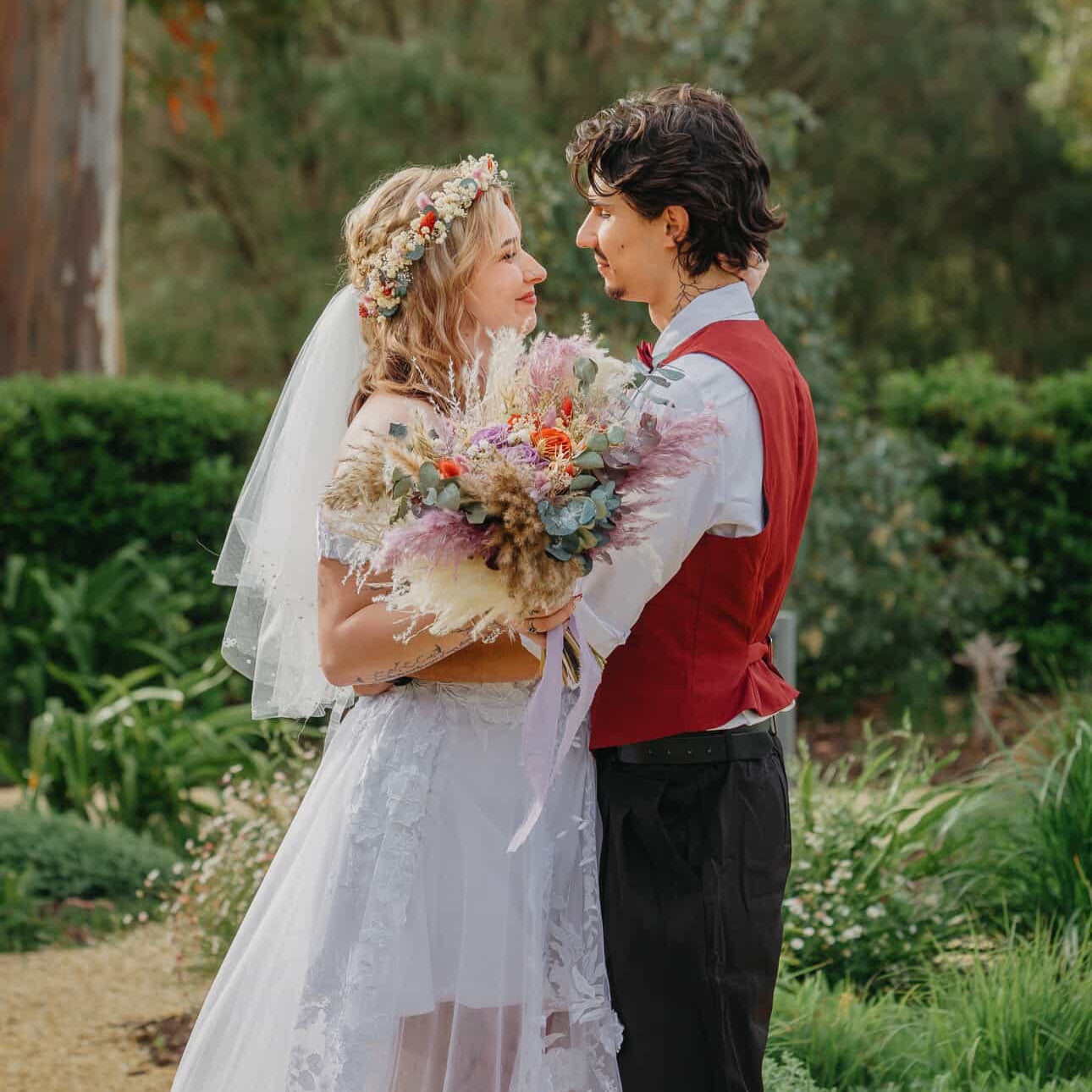 Boho bride holding a large dried flower wedding bouquet while standing with the groom in a lush Australian garden setting.