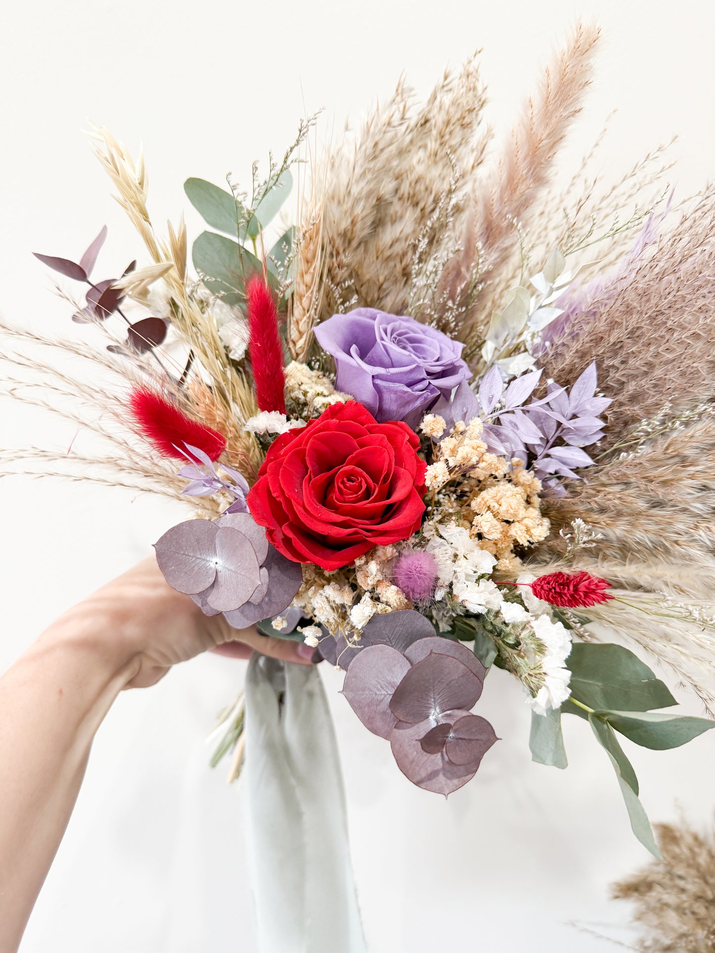 Bouquet of flowers with red roses, purple and pink flowers, and pampas grass held by a person.