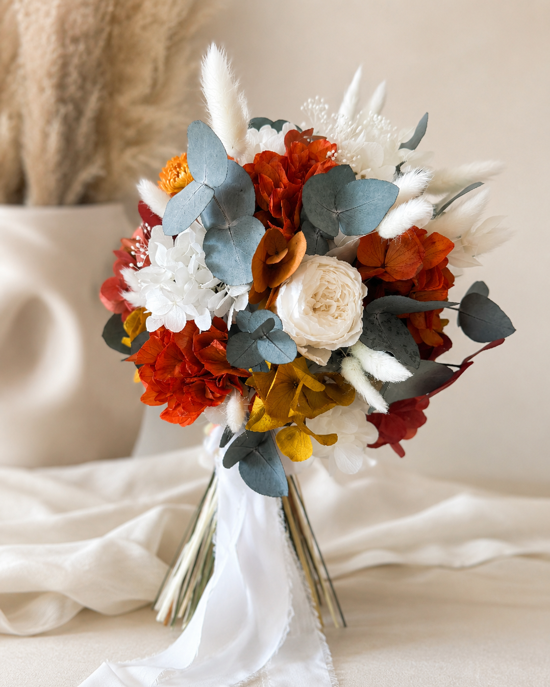 terracotta and burnt orange dried bridal bouquet with a flowing white ribbon, set against a rustic boho background with pampas grass