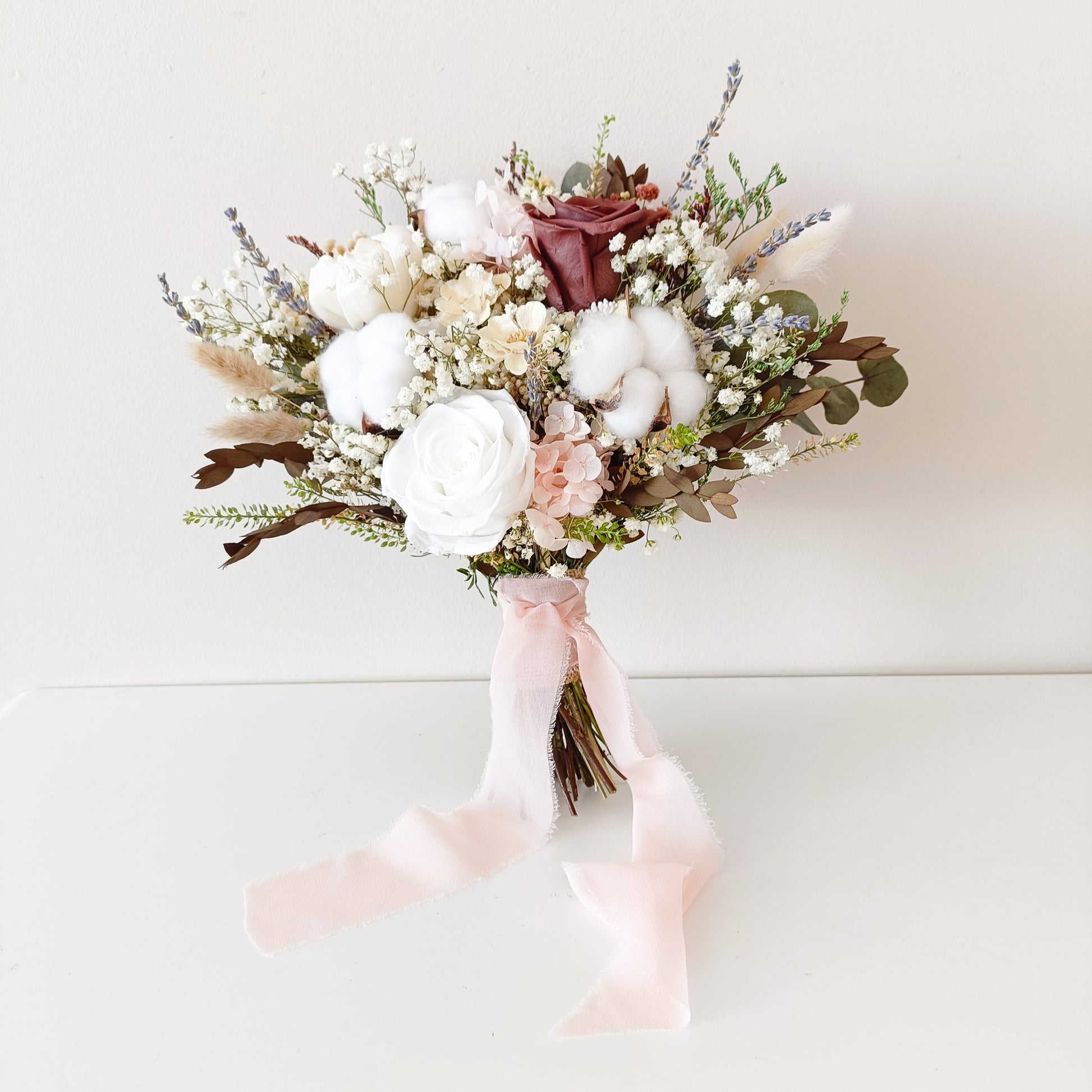 Bridal bouquet made of white sola roses, preserved cotton, and burgundy flowers – wrapped in blush ribbon and styled on a white background.