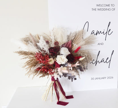 Dried wedding bouquet in burgundy and ivory tones placed in front of a welcome wedding sign – pampas, roses, and preserved florals.