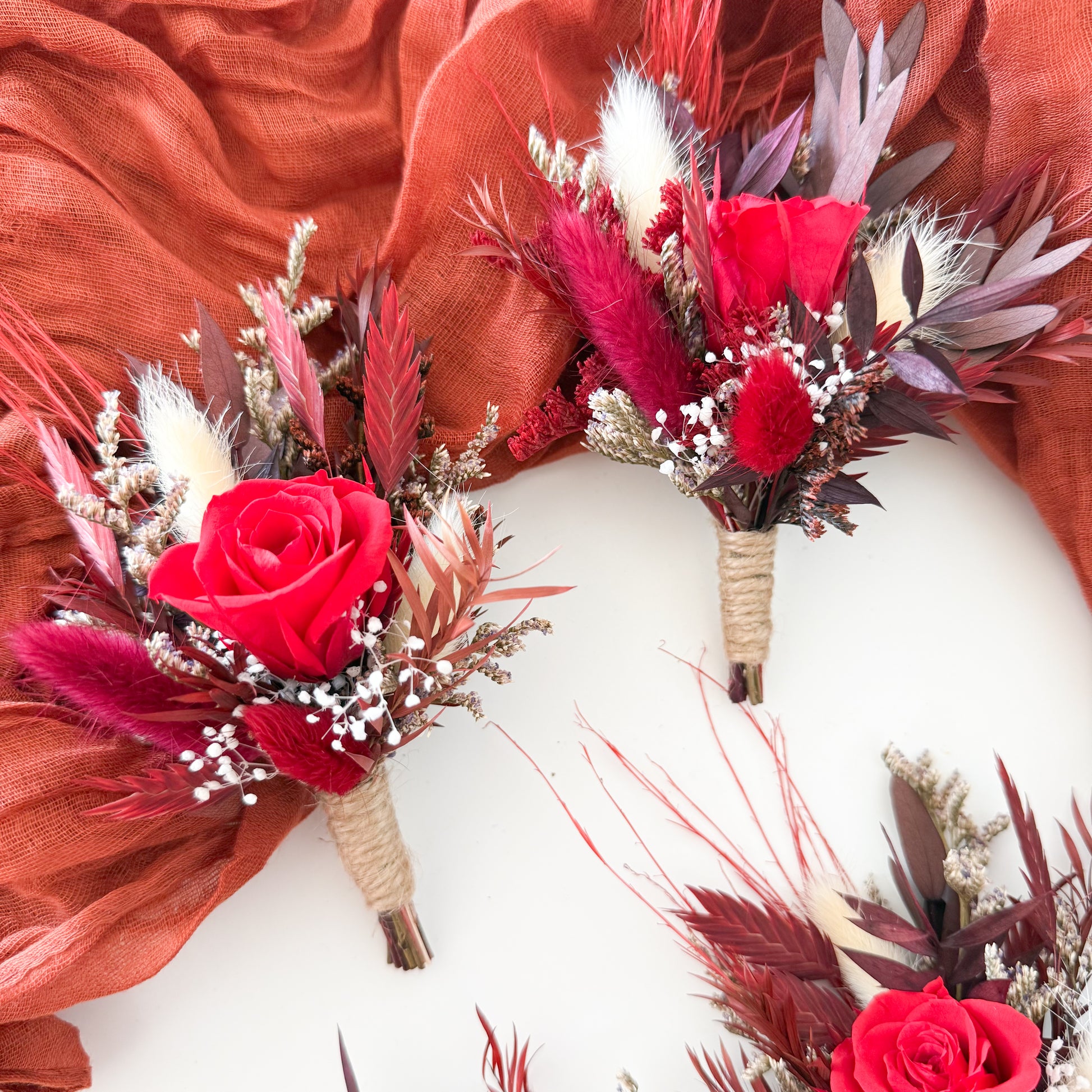 Boho wedding dried flower boutonnieres in red and burgundy tones, sola wood roses with rustic wrap, handmade groomsmen accessories for rustic and bohemian weddings.