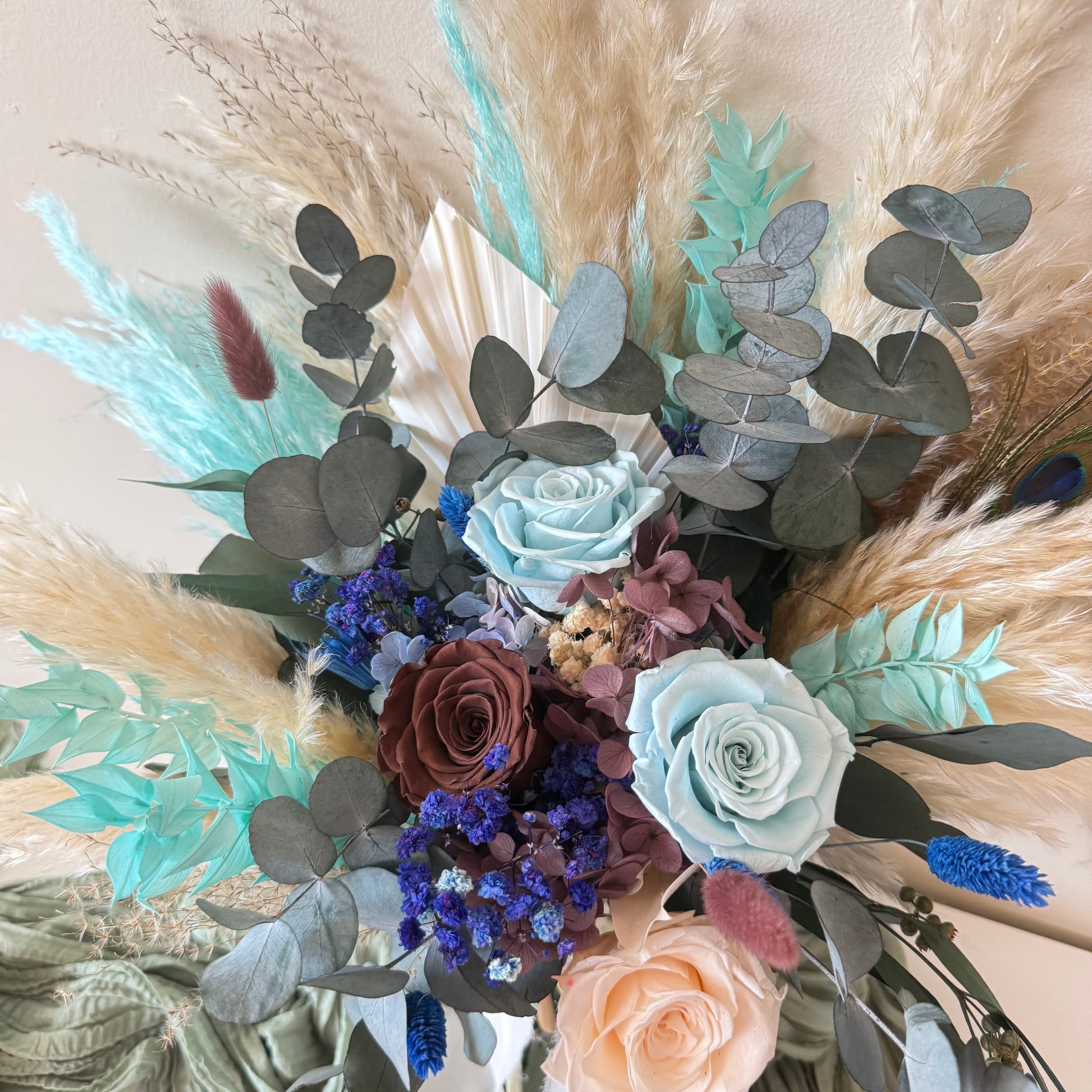 Close-up of teal and navy blue preserved flowers with eucalyptus leaves and beige sola roses.
