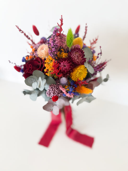 Colorful boho bridal bouquet made with dried flowers in yellow, burgundy, lavender, and peach tones, tied with a red silk ribbon.