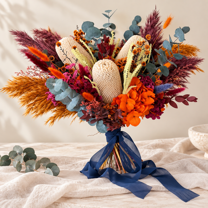 Australian native dried wedding bouquet in a blue vase with cream banksia, orange blooms, burgundy foliage and navy ribbon.
