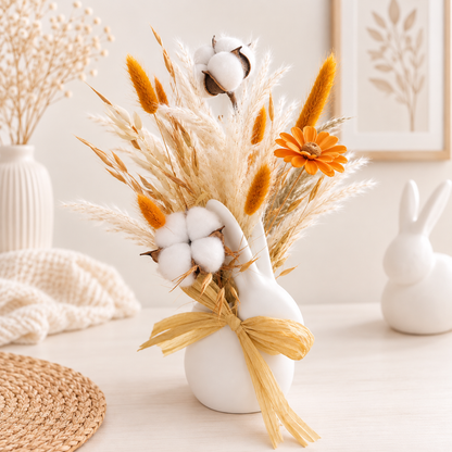 A decorative dried floral arrangement in a small white vase, featuring pampas grass, cotton bolls, orange catkins, and an orange flower, tied with a tan ribbon, styled on a tabletop with home decor props.