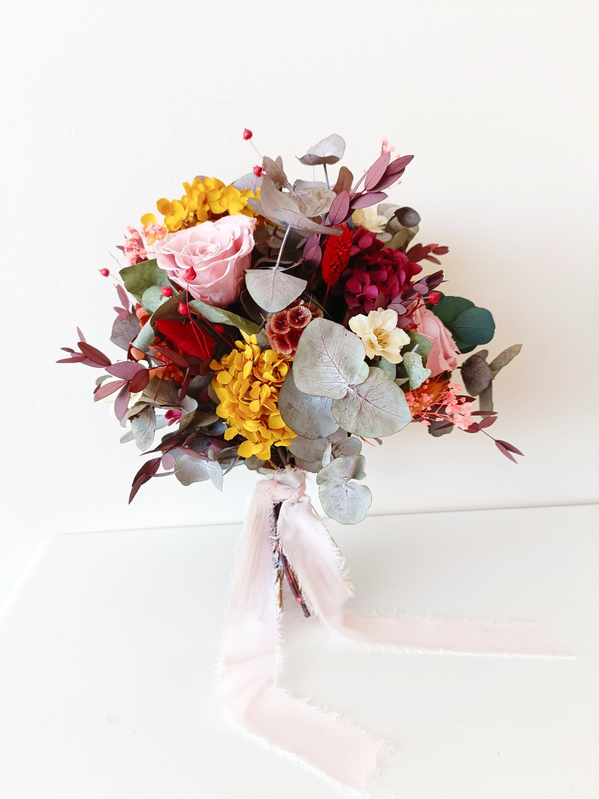 Back angle of dried flower bouquet showcasing eucalyptus leaves, burgundy ruscus and soft preserved rose blooms with long flowing ribbon.