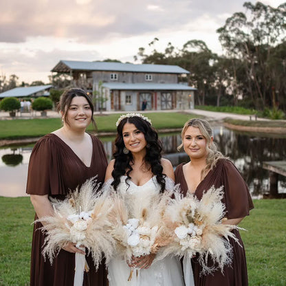 Three women posing outdoors in front of a pond and barn, with the center woman in a white wedding dress and the two women beside her wearing matching brown v-neck dresses. They are holding large beige pampas grass bouquets.