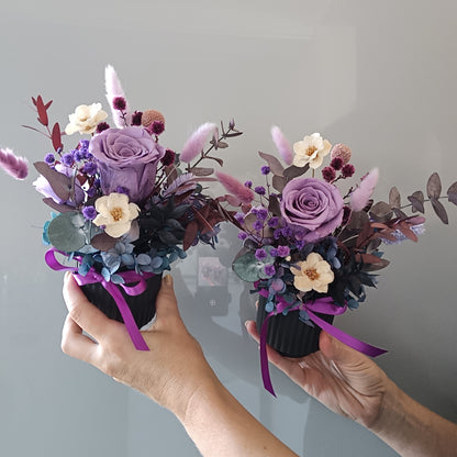 Two hands holding small floral bouquets with purple and white flowers against a gray background