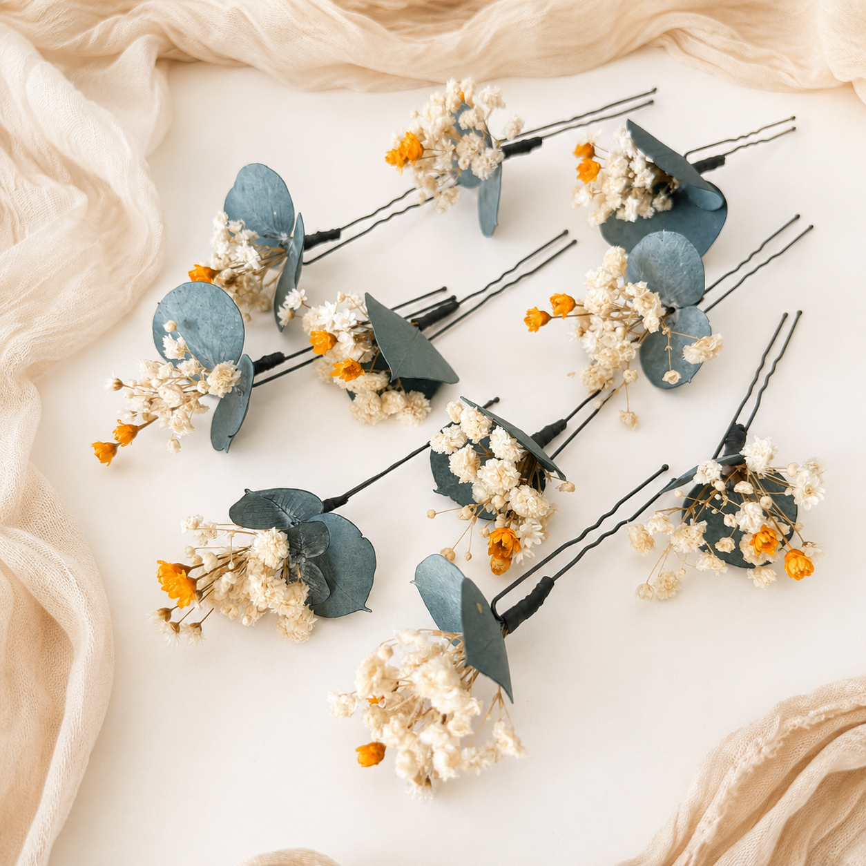 Five dried floral hair pins with dusty blue-green eucalyptus leaves and small orange-yellow flowers, arranged on a cream fabric background.
