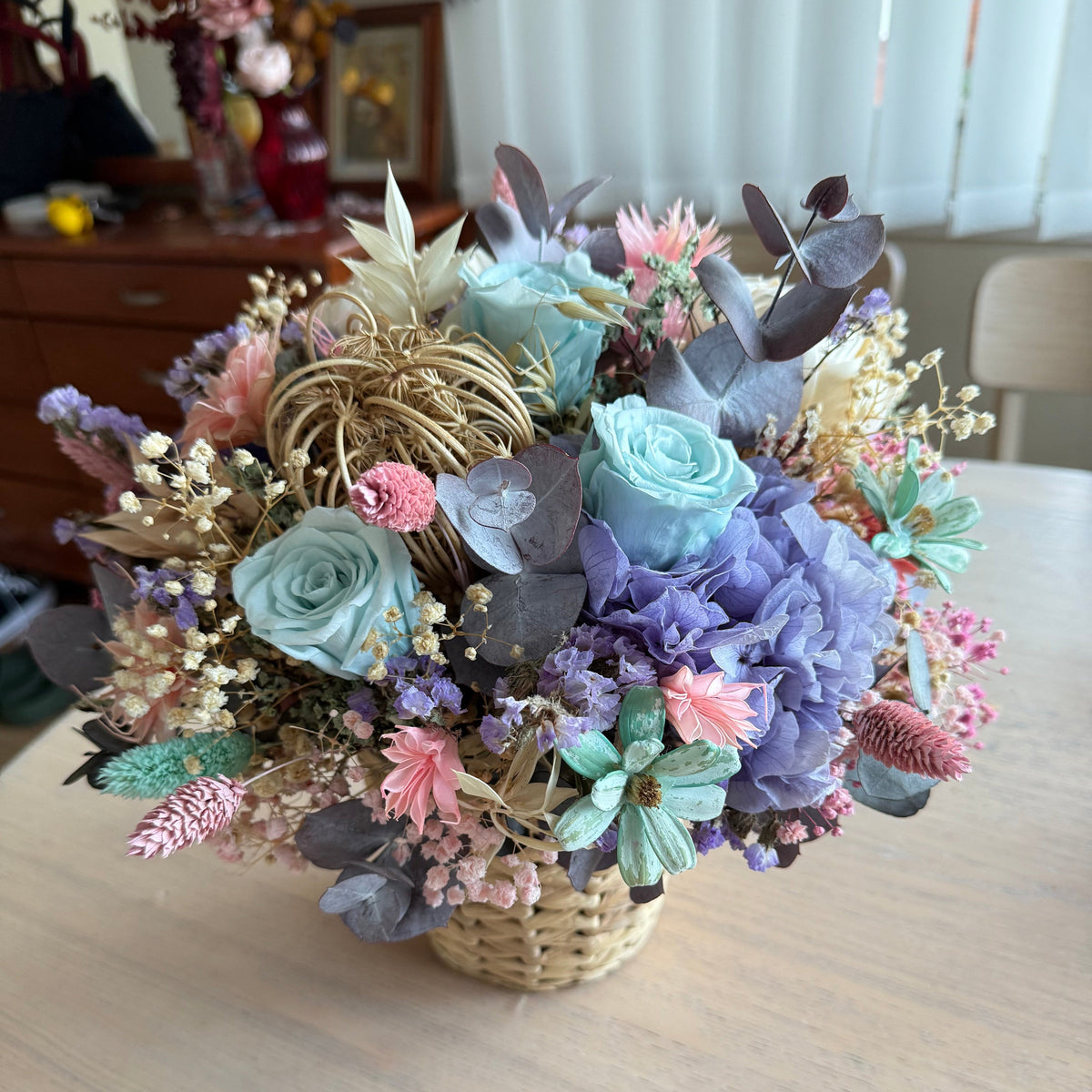 A decorative dried flower arrangement in a woven basket, featuring pastel blue and lavender roses, pink accents, muted green leaves, and dried grasses, displayed on a light wood table.