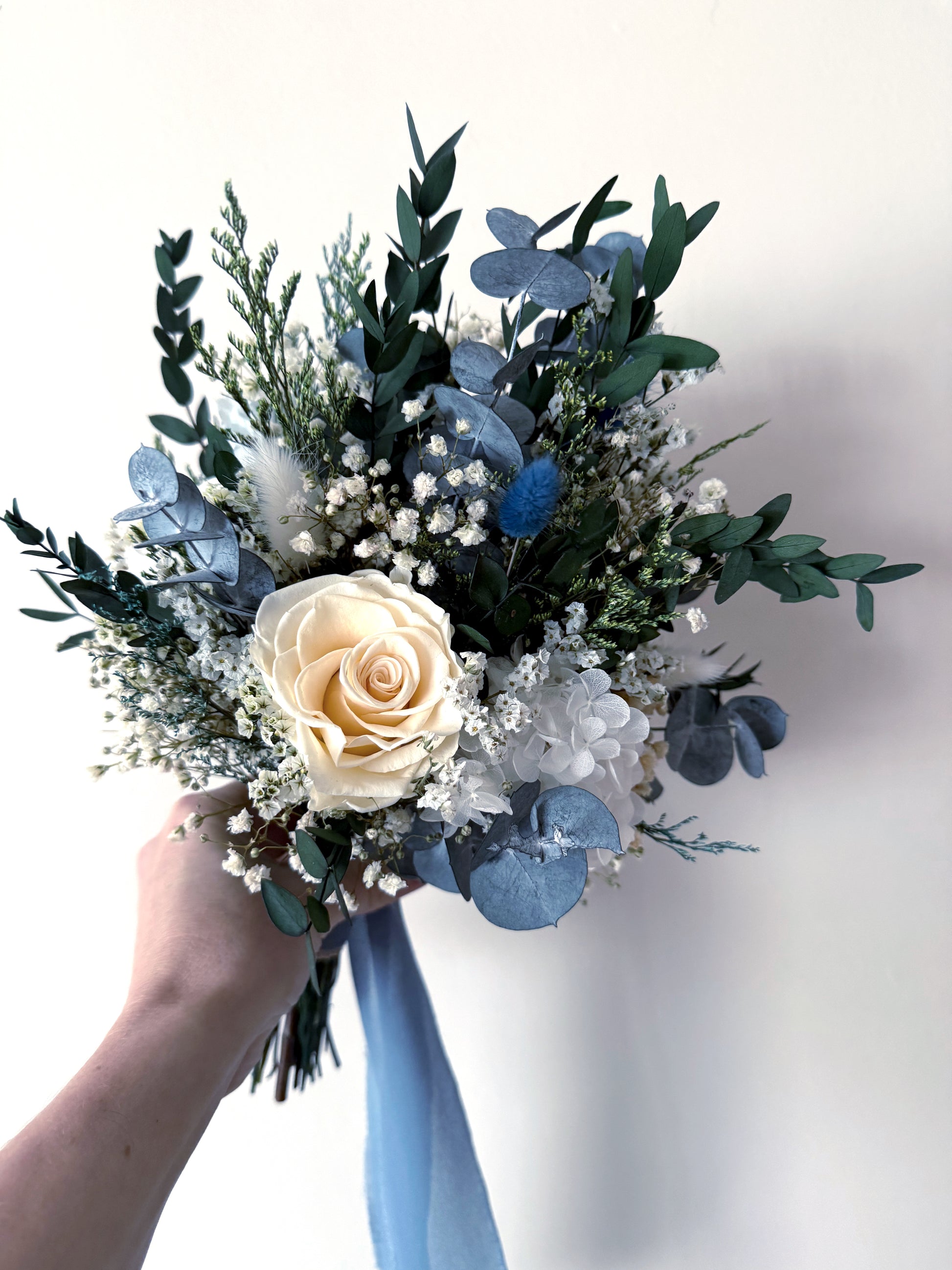 Dried flower bridal bouquet in dusty blue with sola wood roses and natural greenery, finished with flowing blue ribbon.