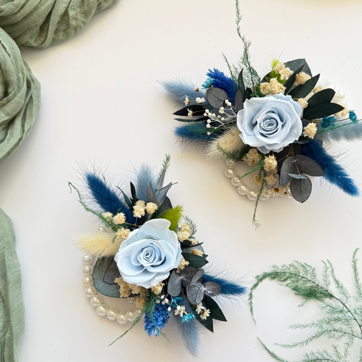 Two floral corsages with blue flowers and greenery on a light background
